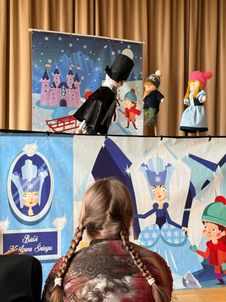 Teatrzyk kukiełkowy na scenie przedstawia postacie z zimowej bajki, na pierwszym planie widać dziecko oglądające przedstawienie.