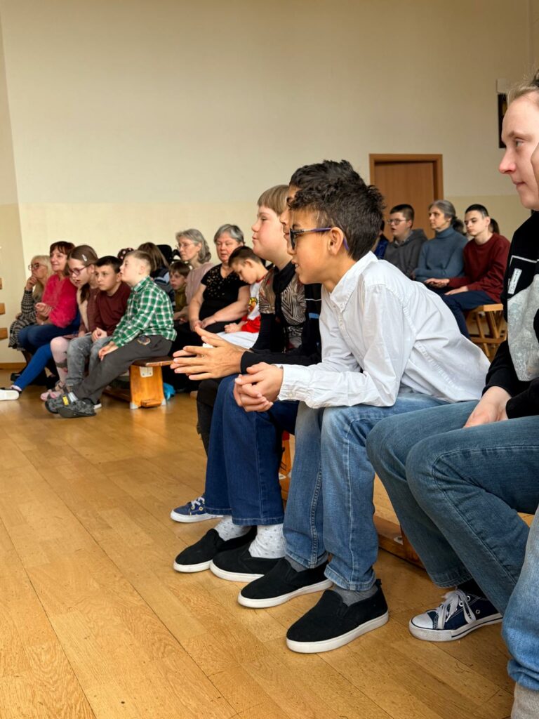 Grupa dorosłych i dzieci siedzi na sali i z uwagą ogląda przedstawienie.