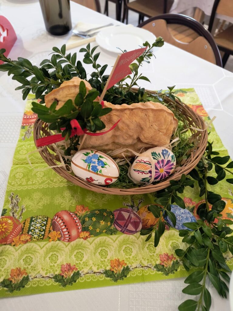 Koszyk wielkanocny z Barankiem, pisankami i zielonymi gałązkami na udekorowanym stole.