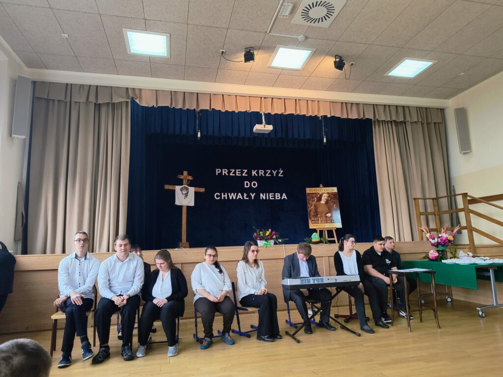 Grupa osób siedzi na scenie w sali, w tle dekoracja religijna z boku koło sceny stół z palmami i jajkami.