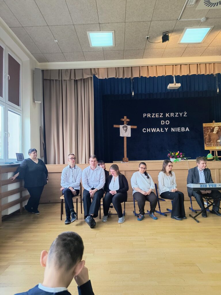 Grupa osób siedzi na scenie w sali, podczas spotkania z okazji życzeń wielkanocnych, w tle dekoracja religijna z krzyżem i napisem.