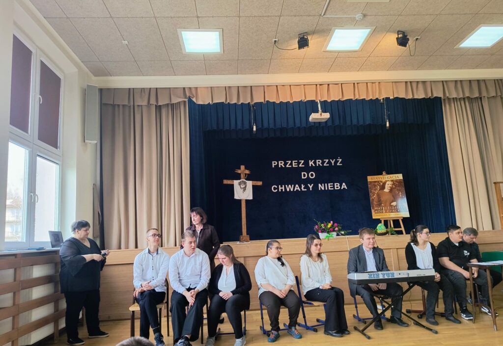 Grupa osób siedzi na scenie w sali, w tle dekoracja religijna z krzyżem i napisem.