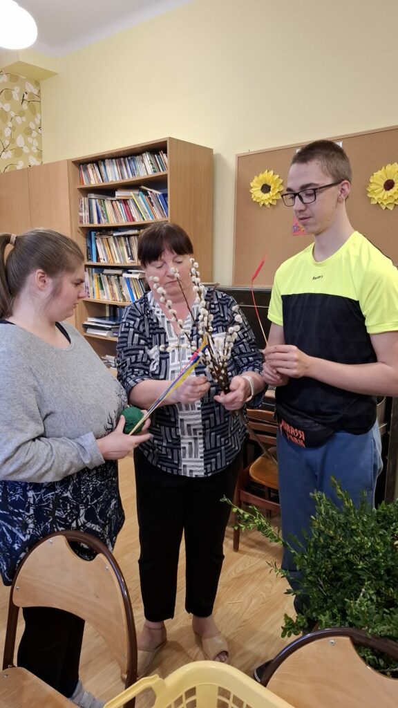 Trzy osoby przygotowują palmę z gałązek i kolorowych elementów.