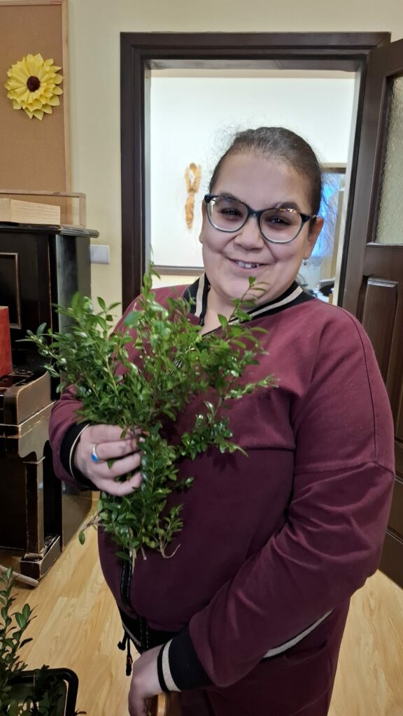 Uśmiechnięta dziewczyna w okularach trzyma zielone gałązki w ręce, stoi w pomieszczeniu.