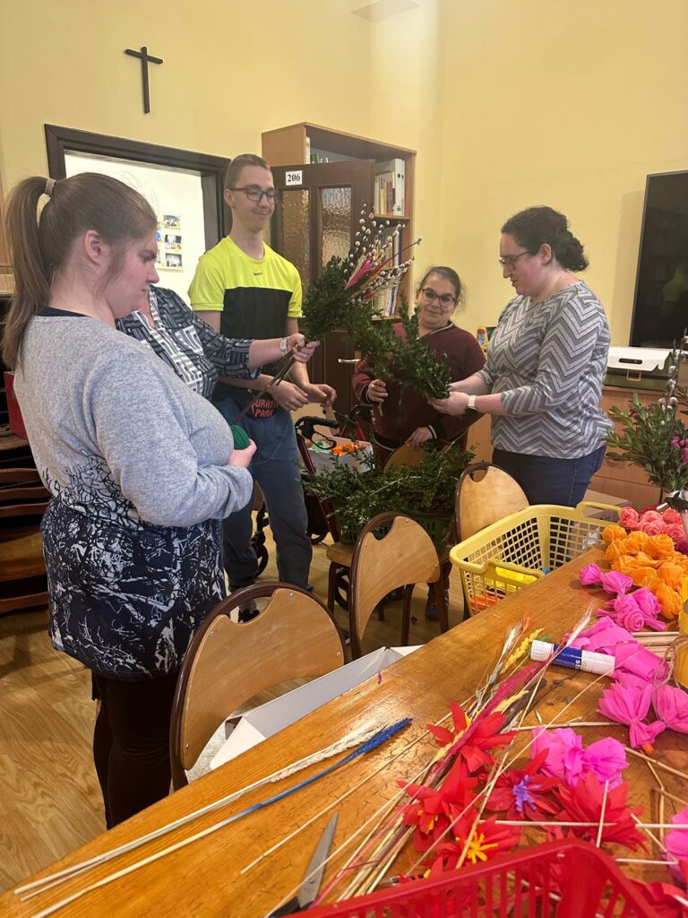 Grupa osób wspólnie tworzy palmę wielkanocną przy stole z materiałami plastycznymi.