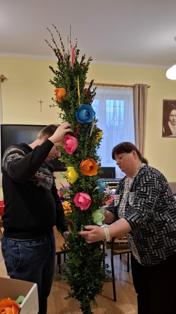 Dwie osoby dekorują palmę kwiatami z papieru.