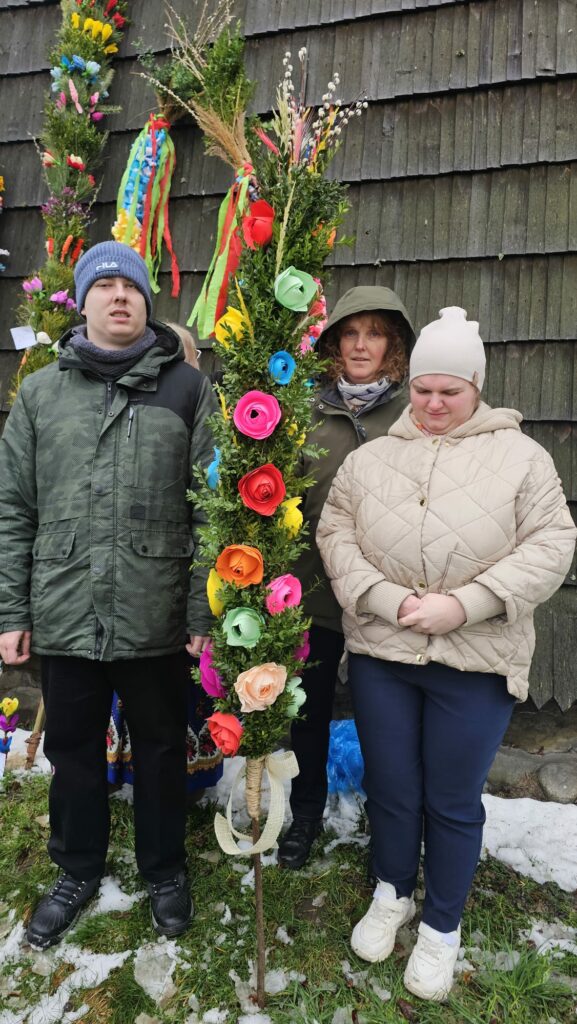 Trzy osoby pozują do zdjęcia na tle starego kościoła z konkursową palmą.