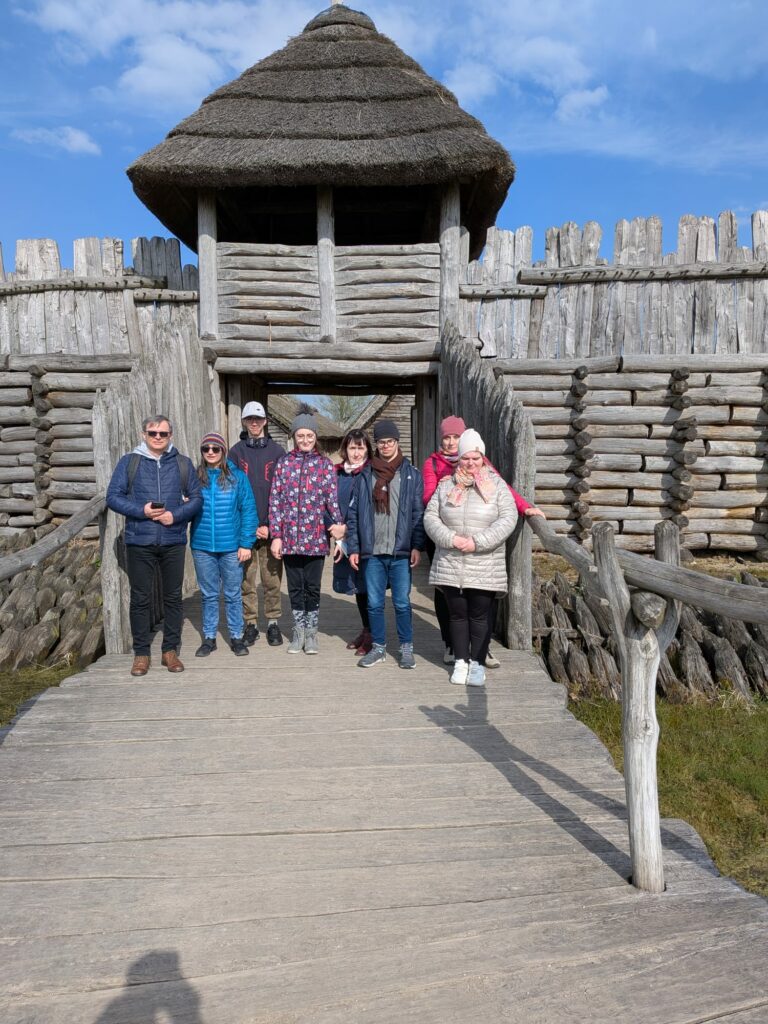 Grupa osób pozuje do zdjęcia przed drewniana bramą i palisadą rekonstrukcji grodu historycznego.