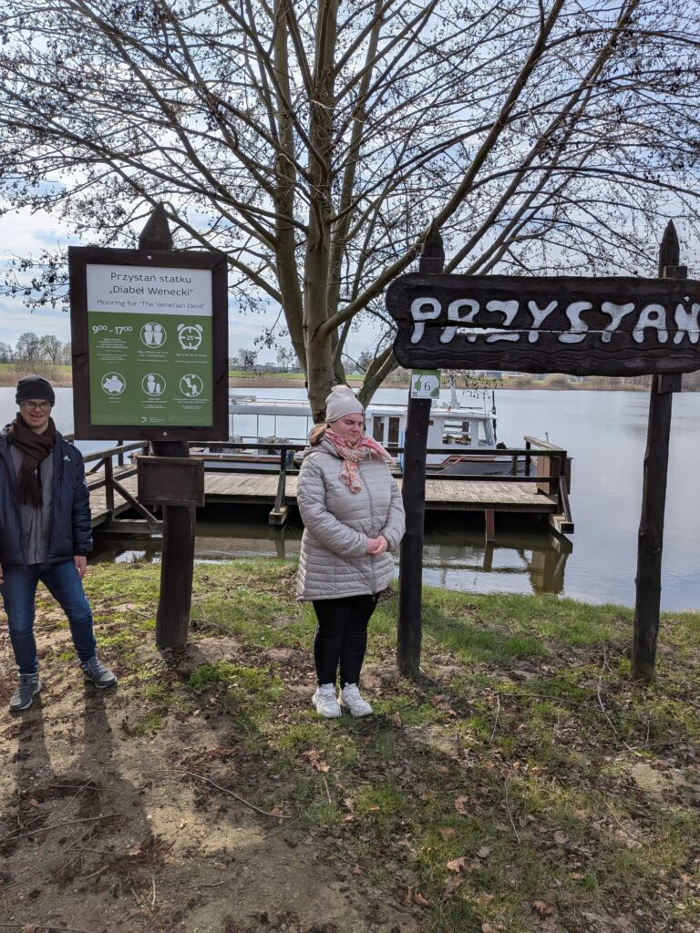 Dwie osoby stoją przy drewnianych tablicach informacyjnych i drogowskazie z napisem "Przystań" nad brzegiem wody.