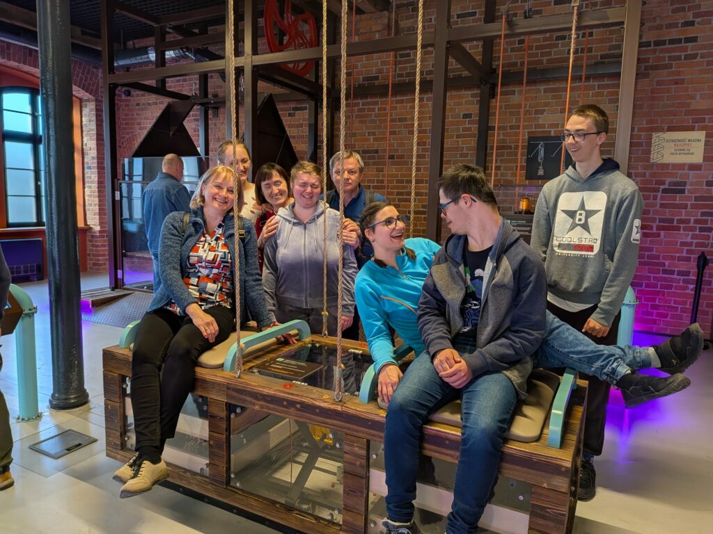 Grupa uśmiechniętych osób siedzi razem na drewnianej konstrukcji wewnątrz muzeum.