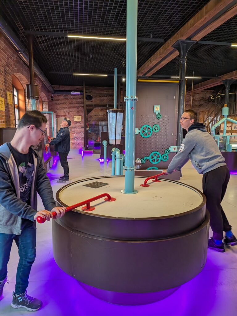 Dwaj młodzi mężczyźni obsługują duże interaktywne urządzenie z korbami.