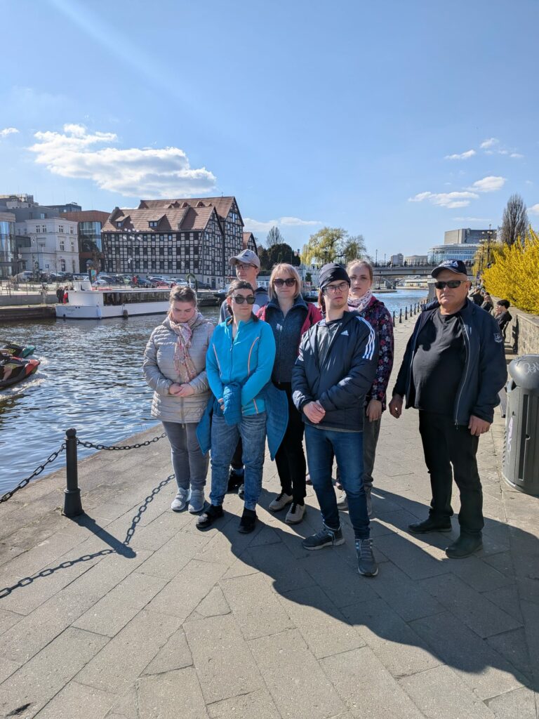 Grupa osób pozuje do zdjęcia na słonecznym bulwarze nad rzeką, w tle widoczna zabytkowa zabudowa miasta.