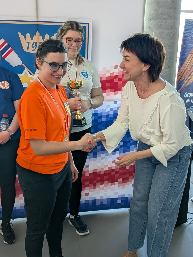 kobieta w pomarańczowej koszulce odbiera gratulacje i uścisk dłoni podczas ceremonii wręczenia nagród.