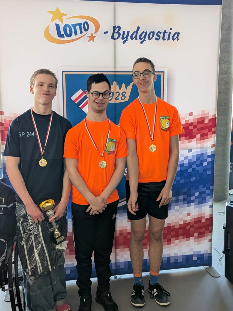 Trzej młodzi mężczyźni w medalach na szyjach pozują na tle ścianki z logo "Lotto - Bydgostia" jeden z nich trzyma puchar.