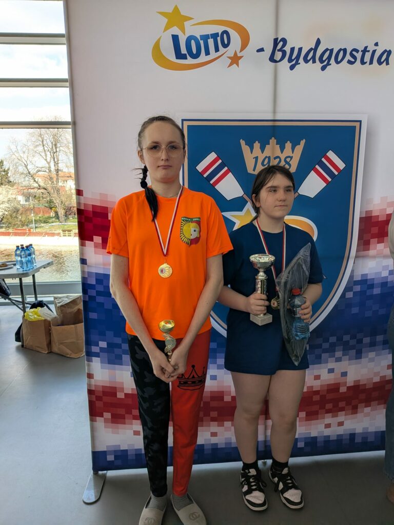 Dwie dziewczyny w strojach sportowych z medalami pozują do zdjęcia.