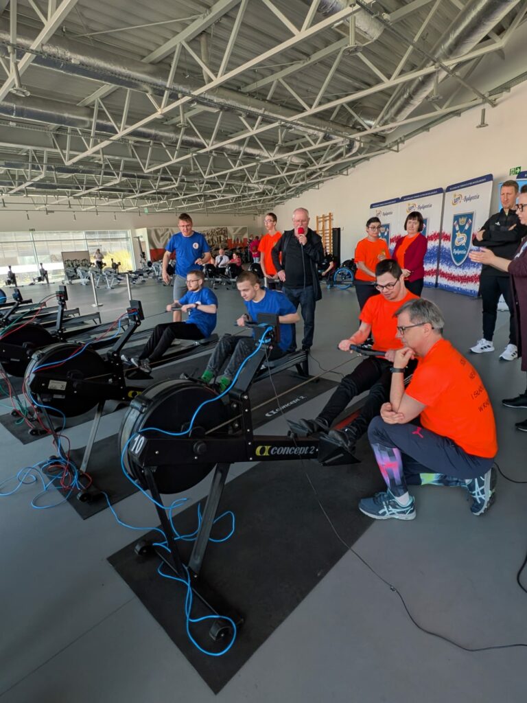 Szerokie ujęcie hali sportowej z wieloma stanowiskami wioślarskimi, przy których trenują zawodnicy pod opieką instruktorów.