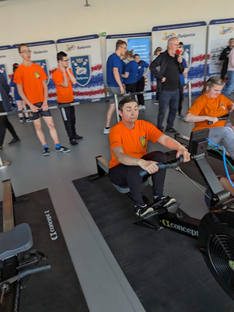 Dwie kobiety podczas zawodów na ergometrach, w tle pozostali uczestnicy.