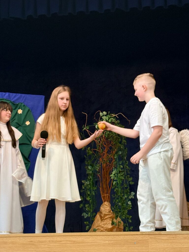 Dziewczyna w białej sukience trzyma w ręce mikrofon i odbiera owoc od chłopca stojącego obok, w tle dekoracja z drzewem.