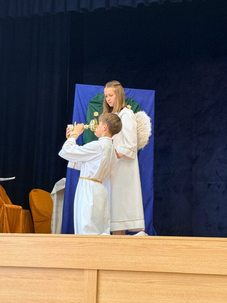 Dzieci w białych strojach aniołów na scenie, jedno podaje kielich drugiemu podczas przedstawienia.