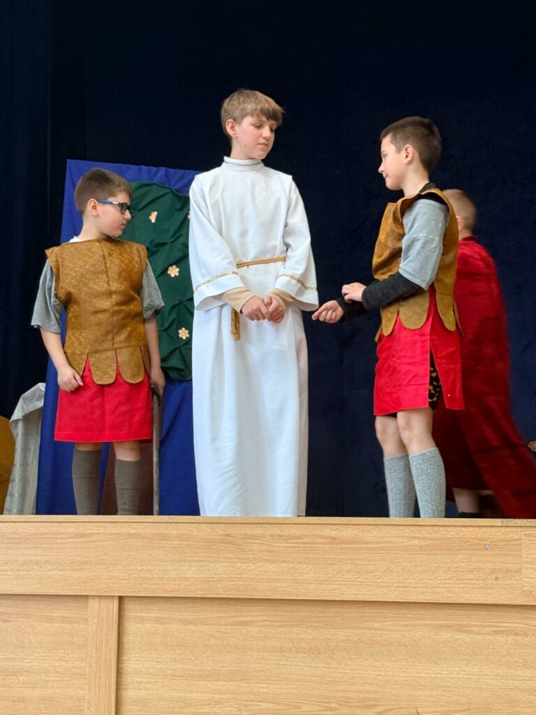 Dzieci w kostiumach historycznych na scenie, chłopiec w białej tunice rozmawia z dwoma innymi chłopcami.