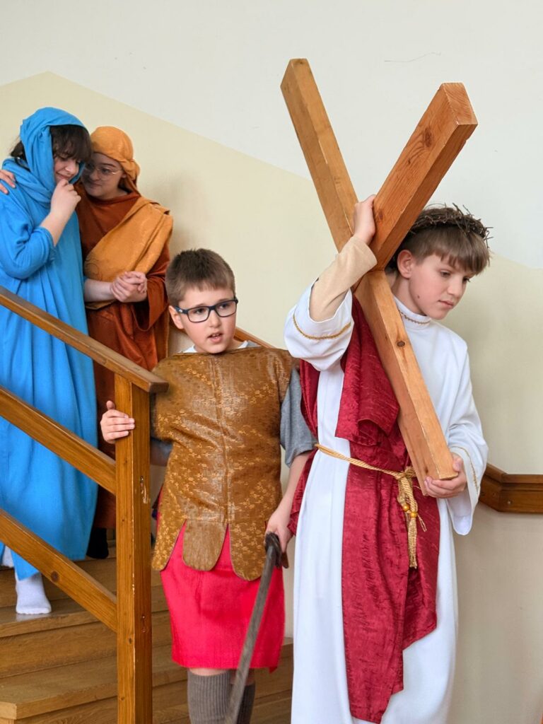 Dzieci w strojach biblijnych na schodach, chłopiec niesie drewniany krzyż, inni towarzyszą mu z tyłu.