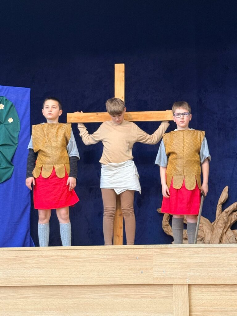 Chłopiec trzyma się drewnianego krzyża, obok obok stoją dwaj inni chłopcy w strojach przypominających rzymskie tuniki.