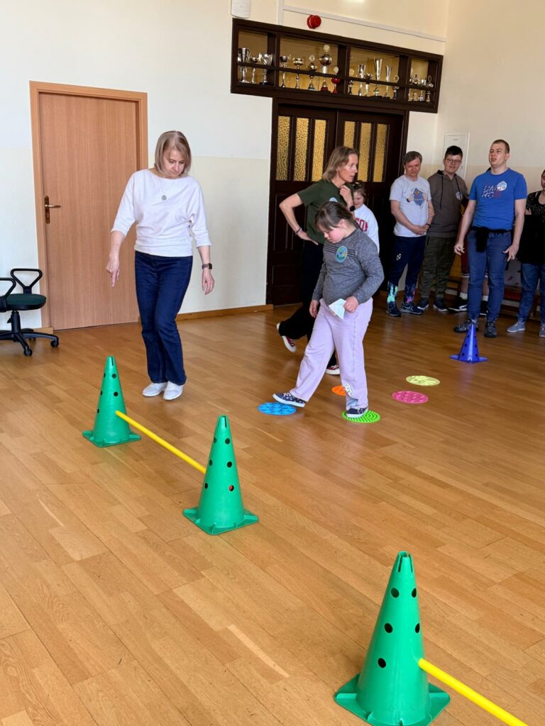 Grupa dzieci i dorosłych na sali gimnastycznej podczas ćwiczeń ruchowych z wykorzystaniem kolorowych pachołków, kamieni sensorycznych i tyczek.