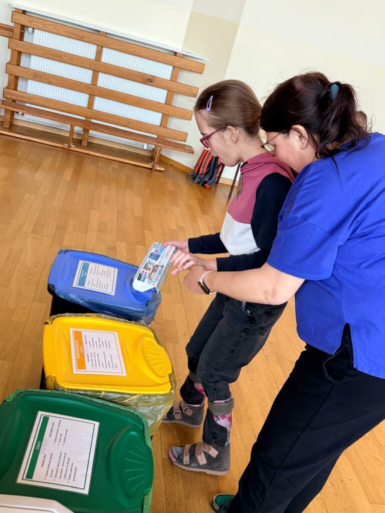 Kobieta pomaga dziewczynce w okularach wrzucić opakowanie do niebieskiego pojemnika na odpady.