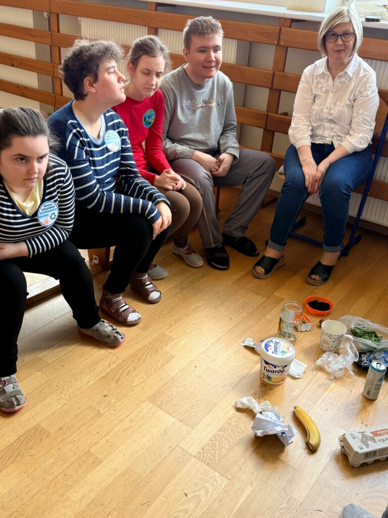 Grupa osób siedzi w kręgu na podłodze leżą różne opakowania po produktach spożywczych.