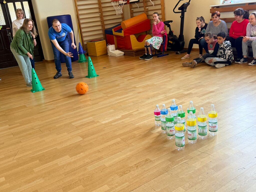 Młody mężczyzna na sali gimnastycznej rzuca piłką do kręgli wykonanych z plastikowych butelek.