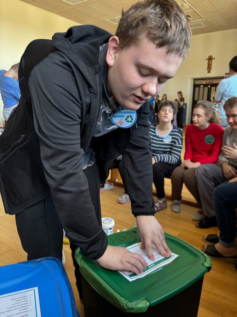 Chłopiec pochyla się nad zielonym koszem na śmieci czytając w Brajlu etykietę. 