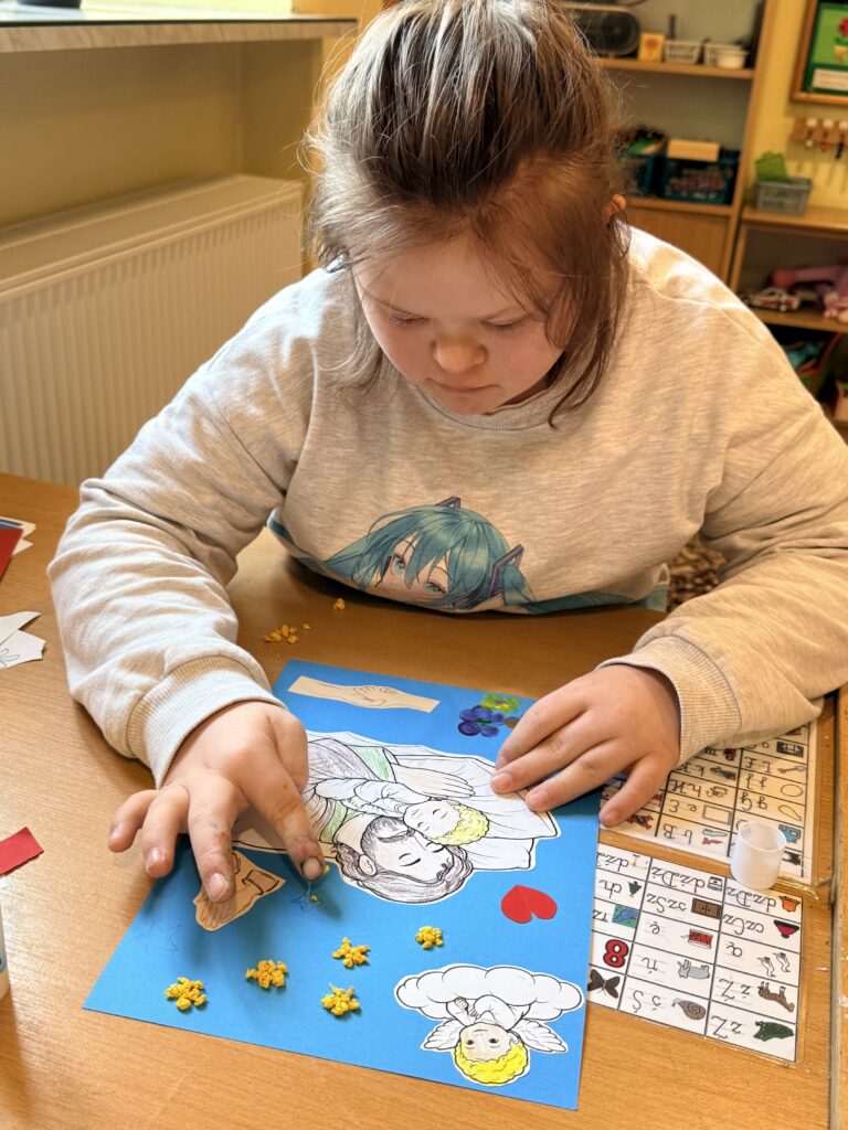 Dziewczynka skupiona na pracy plastycznej przy stole, układa elementy na niebieskiej kartce.