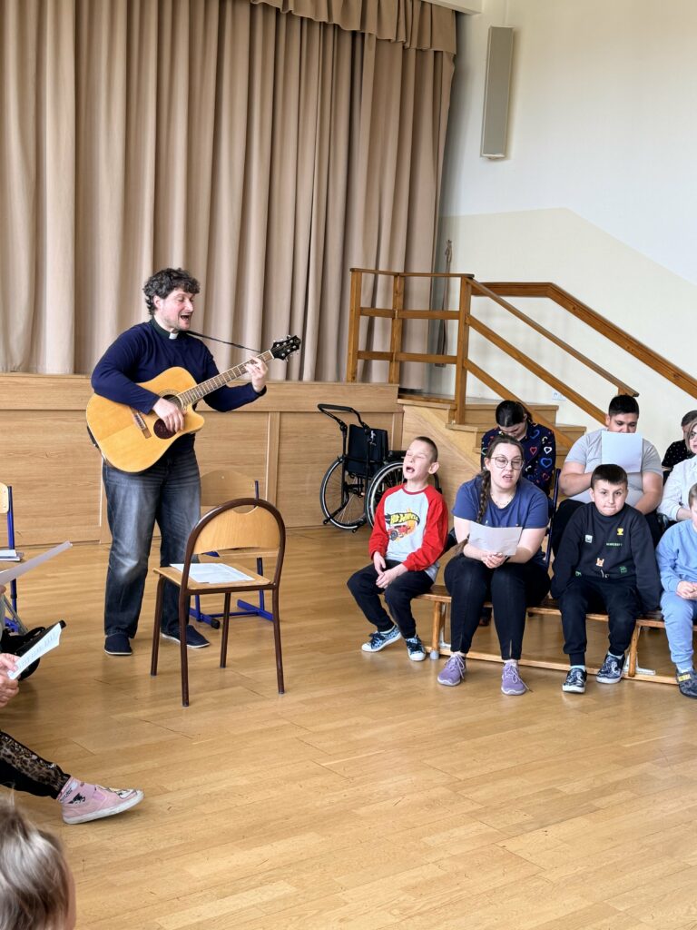 Ksiądz gra na gitarze i śpiewa, a dzieci siedzą obok i uczestniczą w zajęciach.