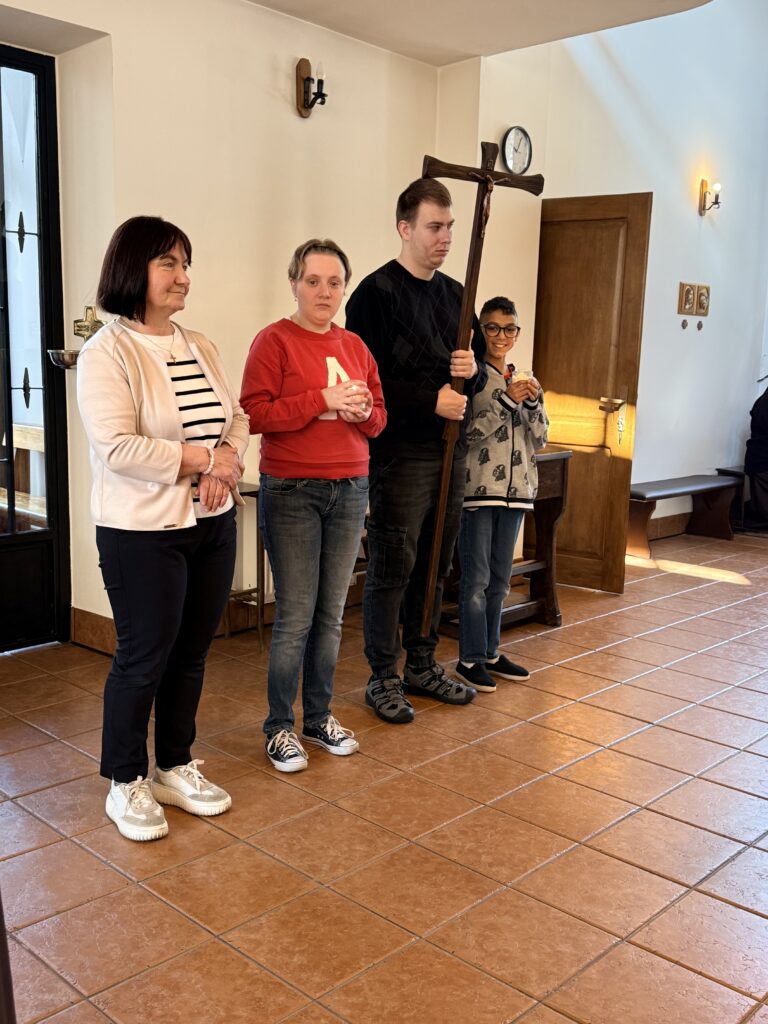 Cztery osoby stoją w kaplicy, trzymają świece i krzyż podczas uroczystości religijnej.