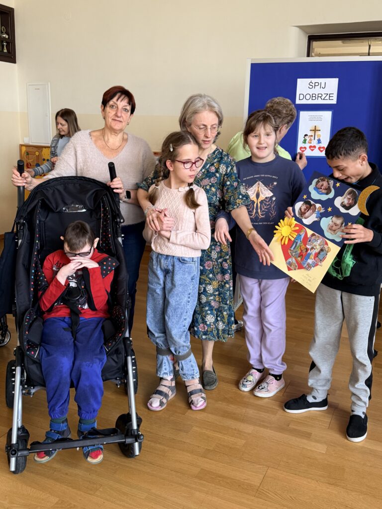 Dwie kobiety pomagają dzieciom, jedno siedzi w wózku, inne trzymają kolorową pracę plastyczną.