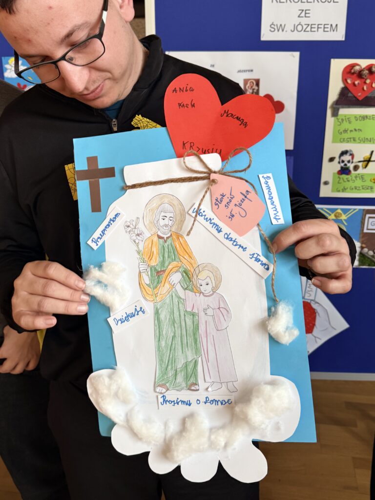 Mężczyzna w okularach trzyma ręcznie wykonaną pracę plastyczną z sercem i rysunkiem o tematyce religijnej.