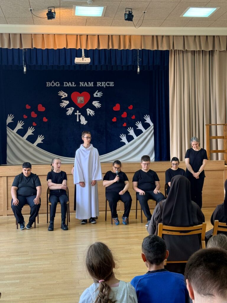 Szersze ujęcie przedstawiające grupę występującą na scenie pod napisem „BÓG DAŁ NAM RĘCE”. Na pierwszym planie widoczny jest tył głów siedzącej publiczności, w tym siostry zakonnej w habicie.