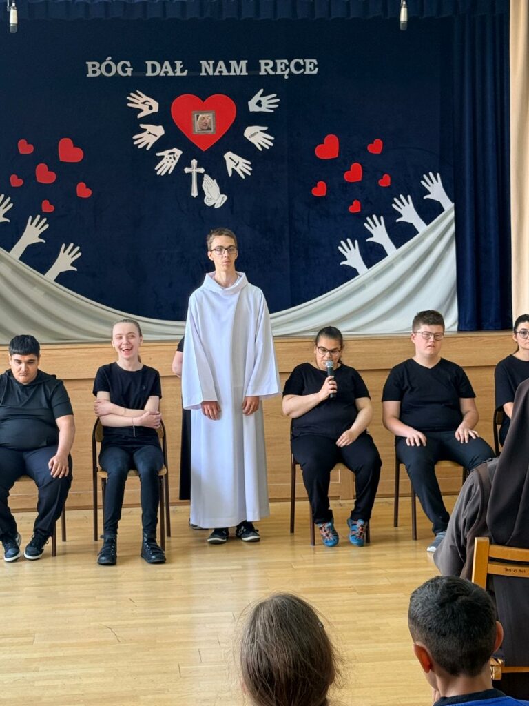Chłopak w długiej białej szacie liturgicznej stoi na środku sceny przed siedzącymi na krzesłach rówieśnikami w czarnych strojach. Jedna z dziewcząt trzyma mikrofon. W tle znajduje się dekoracja z dużym czerwonym sercem