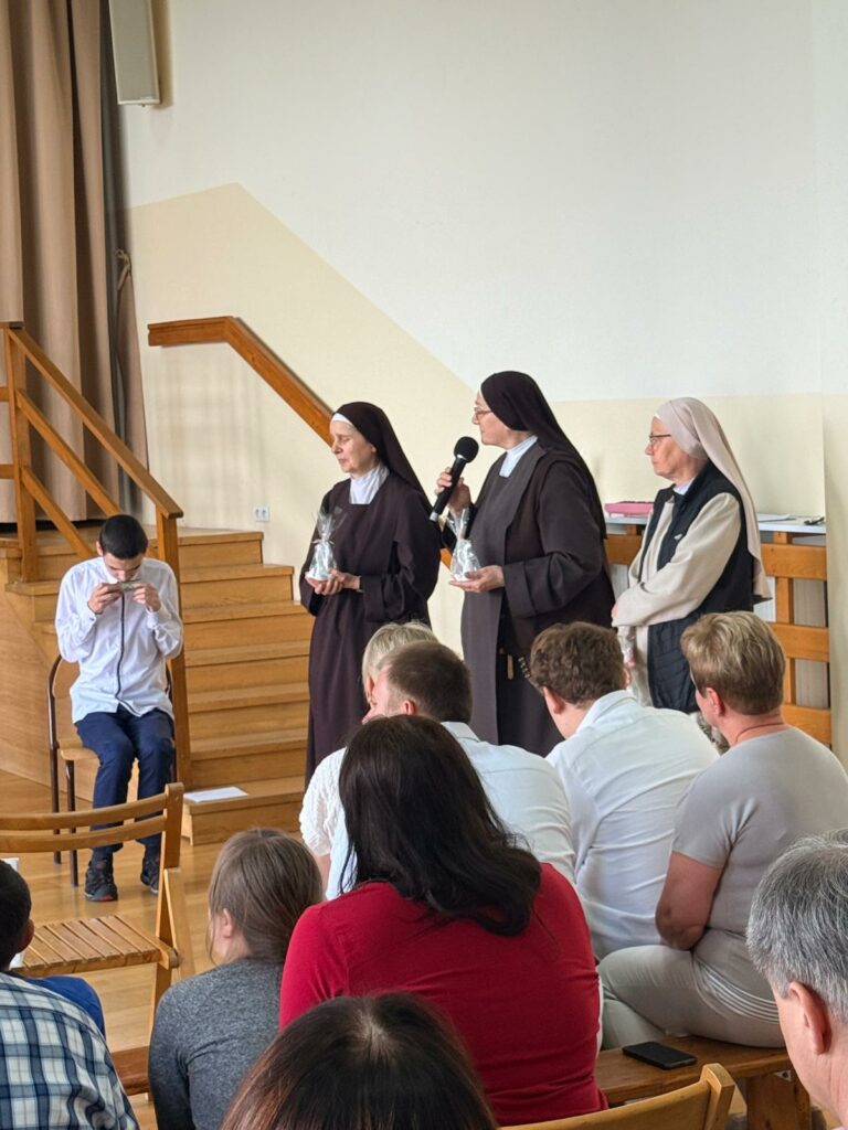 Trzy siostry zakonne stoją obok schodków prowadzących na scenę; jedna z nich mówi do mikrofonu, trzymając upominek. Po lewej stronie siedzi chłopak w białej koszuli, a na pierwszym planie widoczna jest siedząca publiczność.