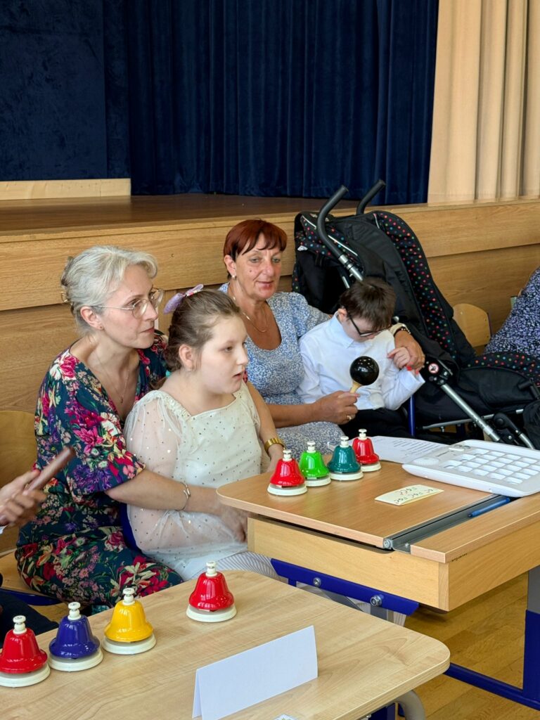 Dziewczynka wspierana przez opiekunkę siedzi przy stoliku z kolorowymi dzwonkami; w tle inne dziecko trzyma marakas.