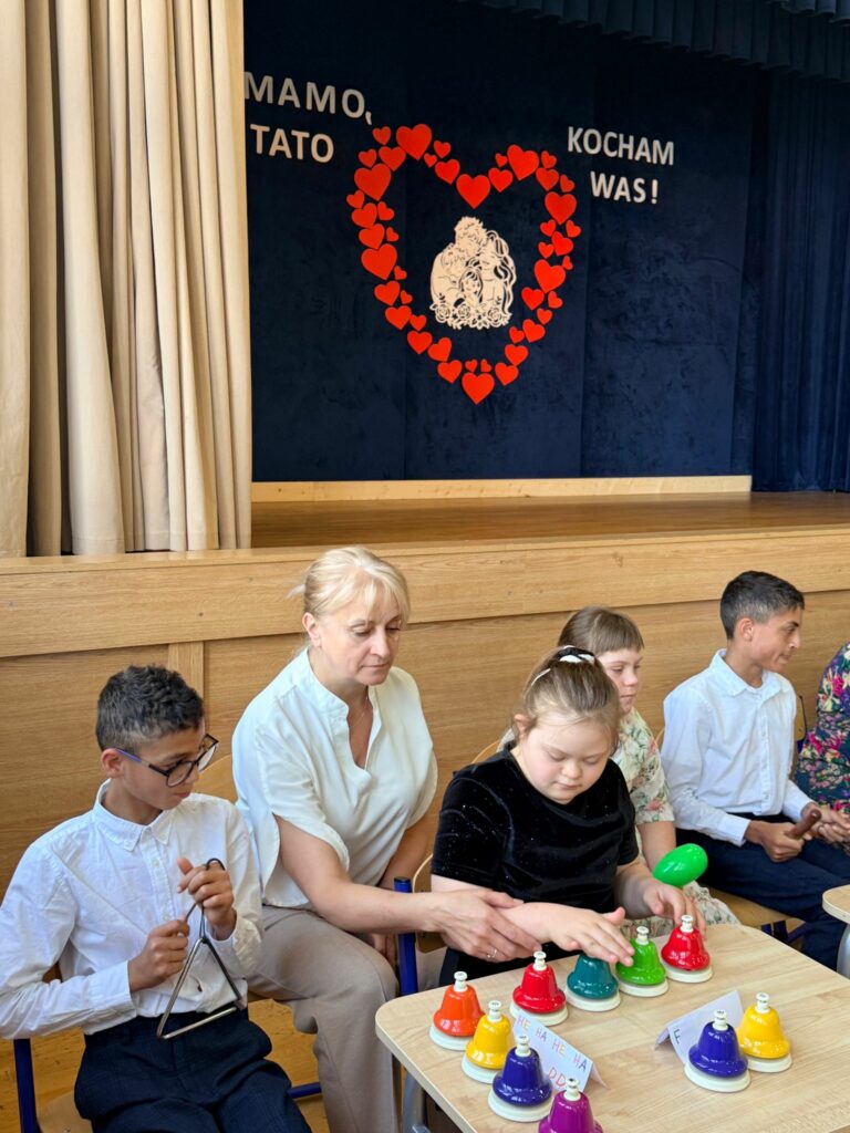 Dziewczynka z zespołem Downa gra na kolorowych dzwonkach biurkowych w asyście nauczycielki; obok chłopiec gra na trójkącie muzycznym. W tle dekoracja z dużym czerwonym sercem.