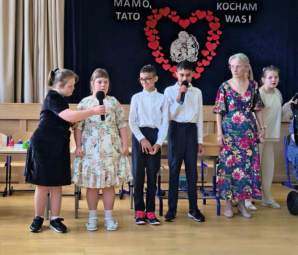Czworo dzieci stoi na scenie z mikrofonami podczas występu, obok nich stoi nauczycielka w kwiecistej sukience. W tle widać dekorację z napisem „Mamo, Tato kocham Was!”.