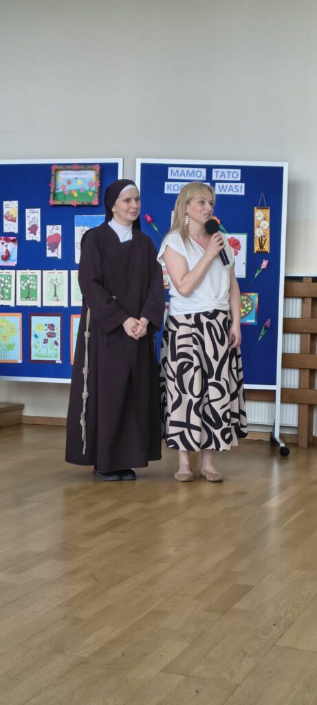 Kobieta z mikrofonem i siostra zakonna stoją na scenie na tle niebieskich tablic z pracami plastycznymi i napisem „Mamo, Tato kocham Was!”.