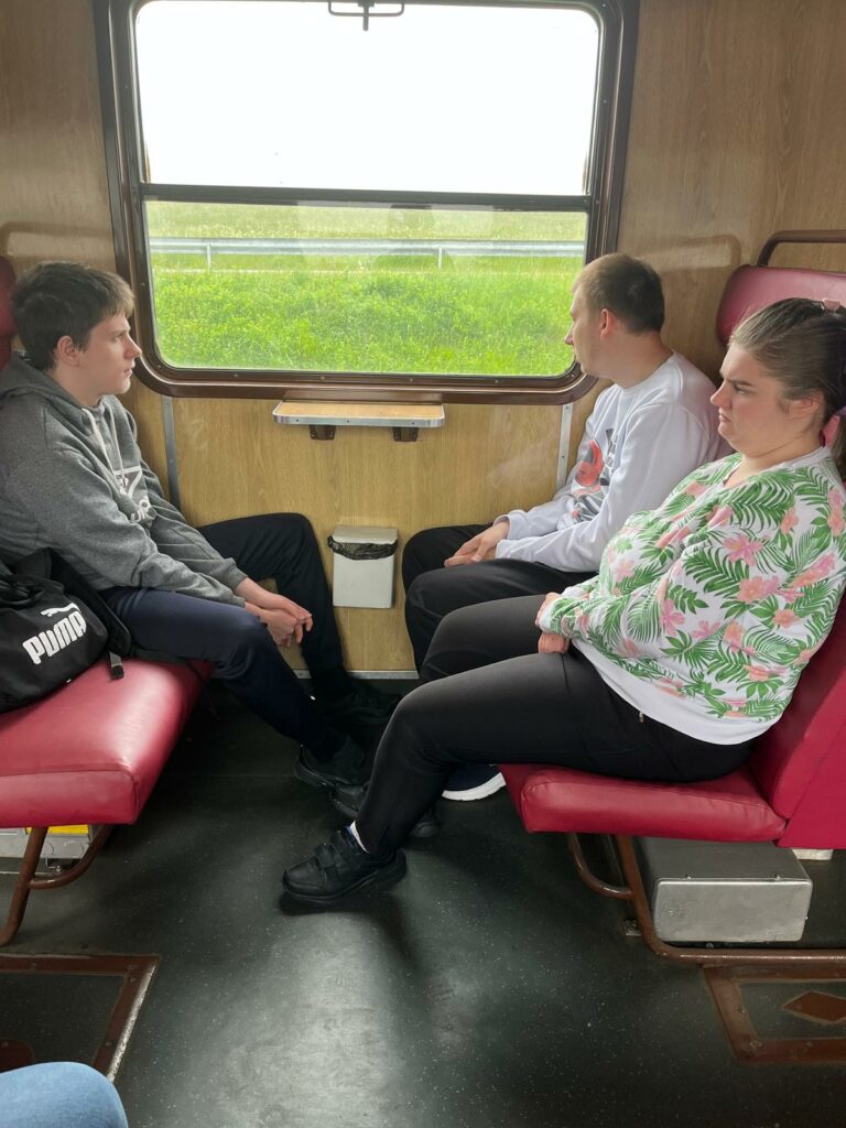 Trzech nastolatków siedzi w przedziale pociągu z bordowymi, skóropodobnymi siedzeniami i drewnianymi panelami na ścianach. Dwoje patrzy prz