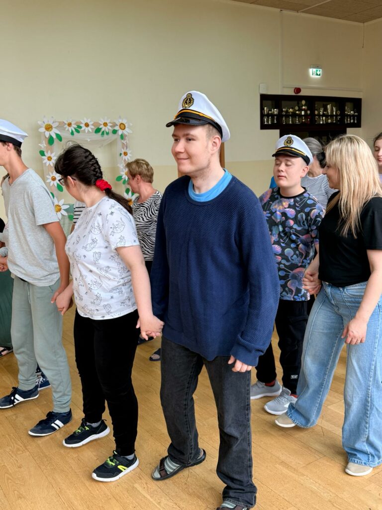 Grupa młodych ludzi, w tym kilku w białych czapkach kapitańskich, tańczy w kółku, trzymając się za ręce, w dużej sali z drewnianą podłogą i widocznymi trofeami w tle.