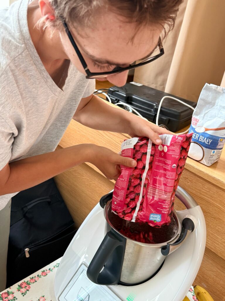 Młody mężczyzna w okularach wsypuje mrożone maliny z foliowego opakowania do misy robota kuchennego Thermomix, stojącej na stole przykrytym obrusem w kwiaty.
