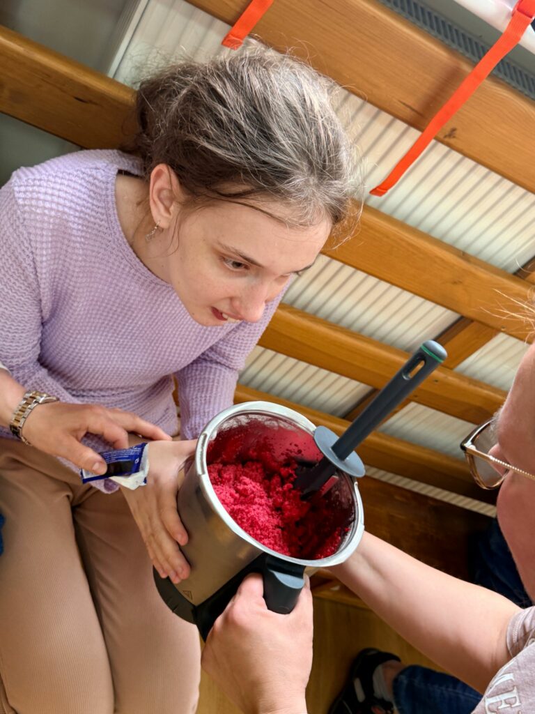 Kobieta w fioletowym swetrze pochyla się nad misą robota Thermomix, trzymaną przez inną osobę, i zagląda do środka na zmiksowaną, czerwoną masę (prawdopodobnie malinową).
