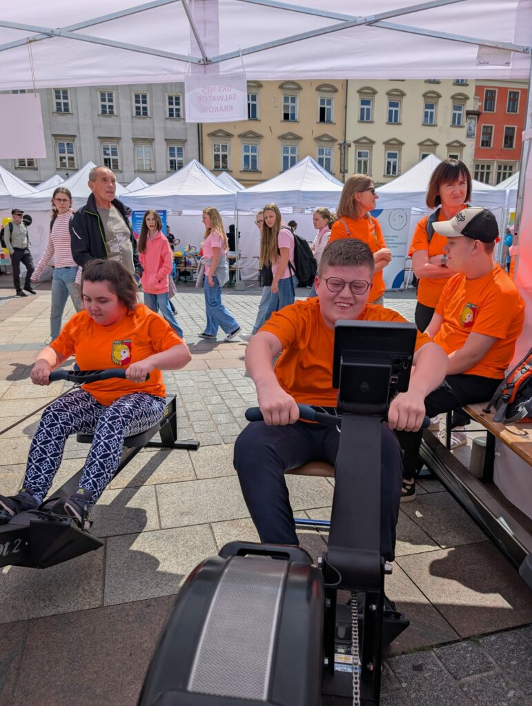 Dwie osoby w pomarańczowych koszulkach korzystają z ergometrów wioślarskich pod namiotem podczas zawodów.