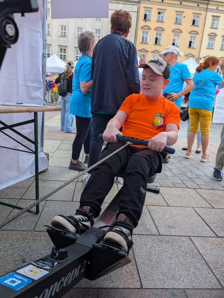 Osoba w pomarańczowej koszulce ćwiczy na ergometrze wioślarskim podczas plenerowego wydarzenia na rynku.