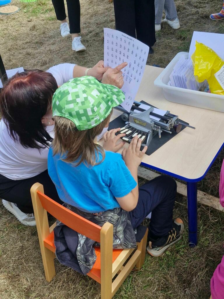 Dziecko w zielonej czapce z daszkiem siedzi przy małym stoliku i uczy się pisać na maszynie brajlowskiej. Dorosła osoba obok wskazuje palcem na arkusz z alfabetem Braille'a.